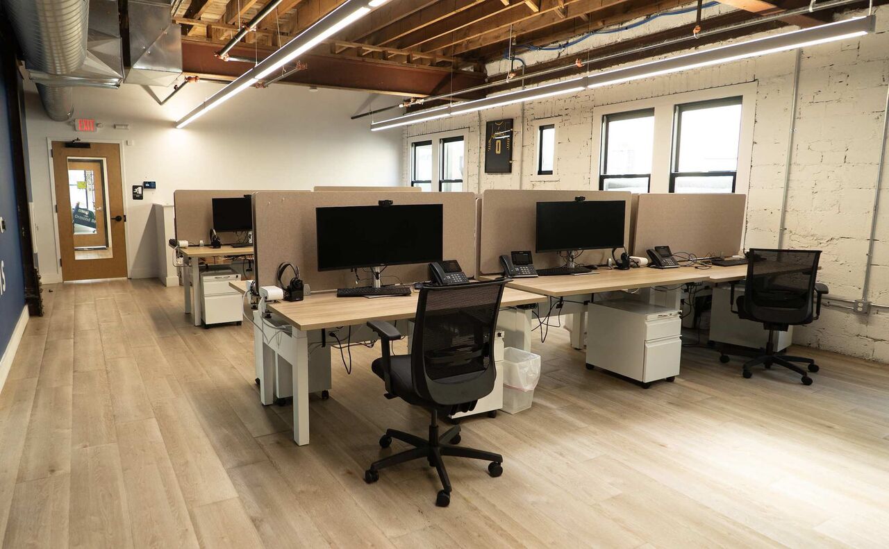 The open desk area at The Node: sit-stand desks with monitors and desk phones under exposed timber beams and white-painted brick