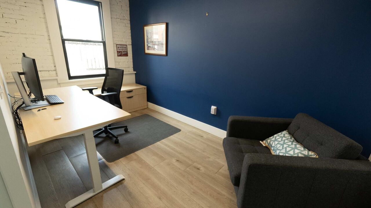 A private office at The Node: a sit-stand desk with a monitor by the window, a navy accent wall and a sofa